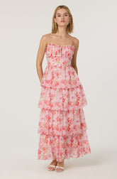 A woman is wearing a sleeveless, tiered maxi dress with thin spaghetti straps. The dress features a ruffled bodice with a zipper in the front, and it has a pink floral pattern with shades of pink, peach, and coral on a sheer fabric. The dress has multiple layered sections, creating a flowing, bohemian look. She is also wearing light-colored open-toe sandals.